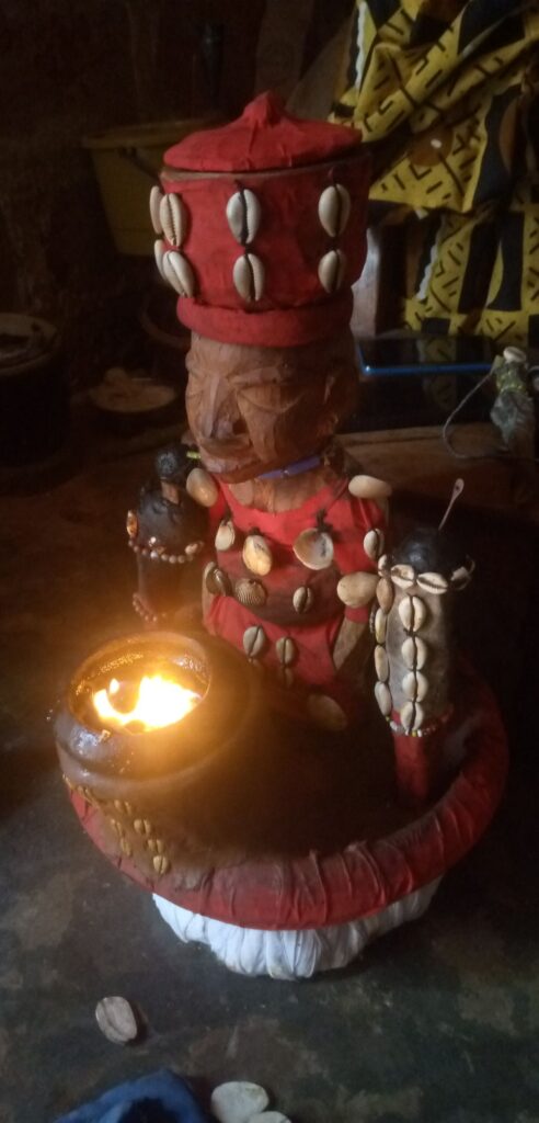 Colorful tiki statue adorned with shells, wearing red clothing, holding a fire bowl in a dimly lit room or yard.